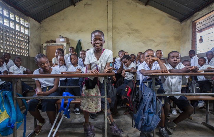 A smiling child is leaning against a desk in a classroom, writing with a pen, and surrounded by more than twenty peers. Democratic Republic of Congo (DRC). Photo credit: HI