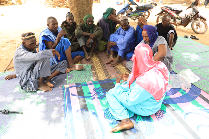 This image shows Aminata Ongoiba, President of a network of community-based organizations called Collectif des déplacés des 7 sites de la commune urbaine. Through daily monitoring and community dialogues, she advocates for quality education for displaced children and supports their continued participation in school. She is shown seated on the ground, engaging in dialogue with male representatives. 