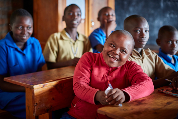 A student sits in a classroom, smiling pleasantly and surrounded by five classmates. There is a blackboard in the background. Rwanda. 