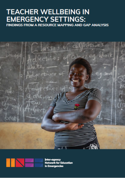 A female teacher in Uganda stands with folded arms in front of a blackboard. She is smiling.